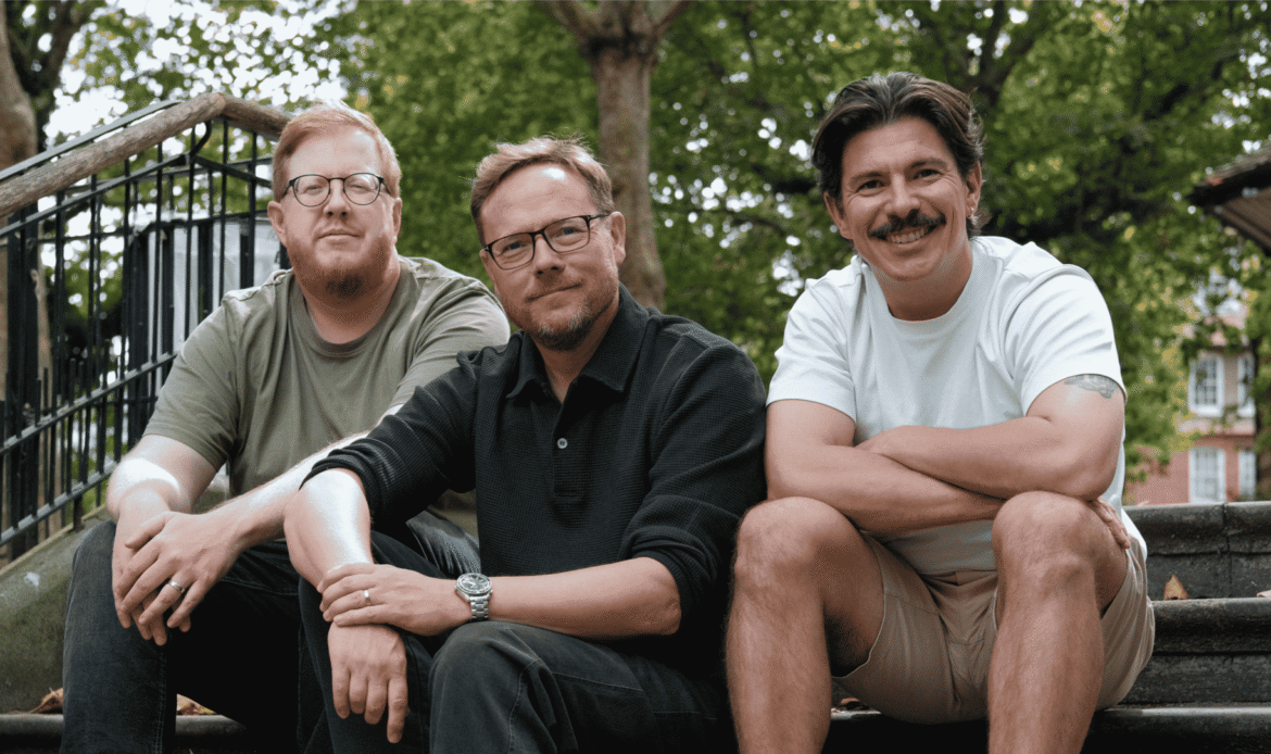 Craig, Andy and Mike are sat on steps in a public park, smiling at the camera.